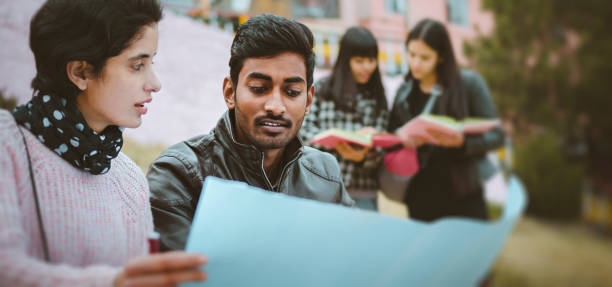 Outdoor panoramic image of happy, multi ethnic, Asian, Indian young adult male and female college student friends discussing project together on chart paper in college campus at day time. Other students are standing in the background. Shoot location Thodo Ground, Solan, (a city in mountain) Himachal Pradesh, India. The panoramic image is also suitable for banner because of more copy space.